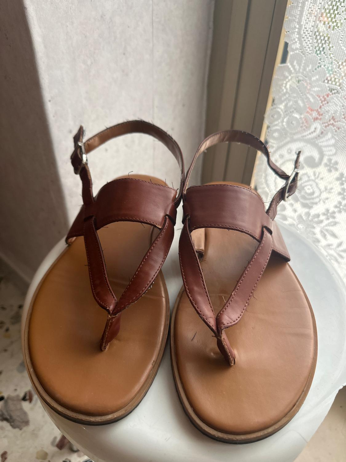 Brown leather sandals with buckles, placed on a white table next to a window with a lace curtain.