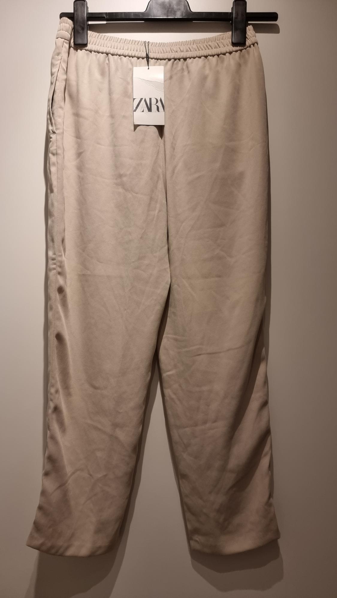 Beige trousers hung on a black hanger with a visible "ZARA" label. Plain white background.