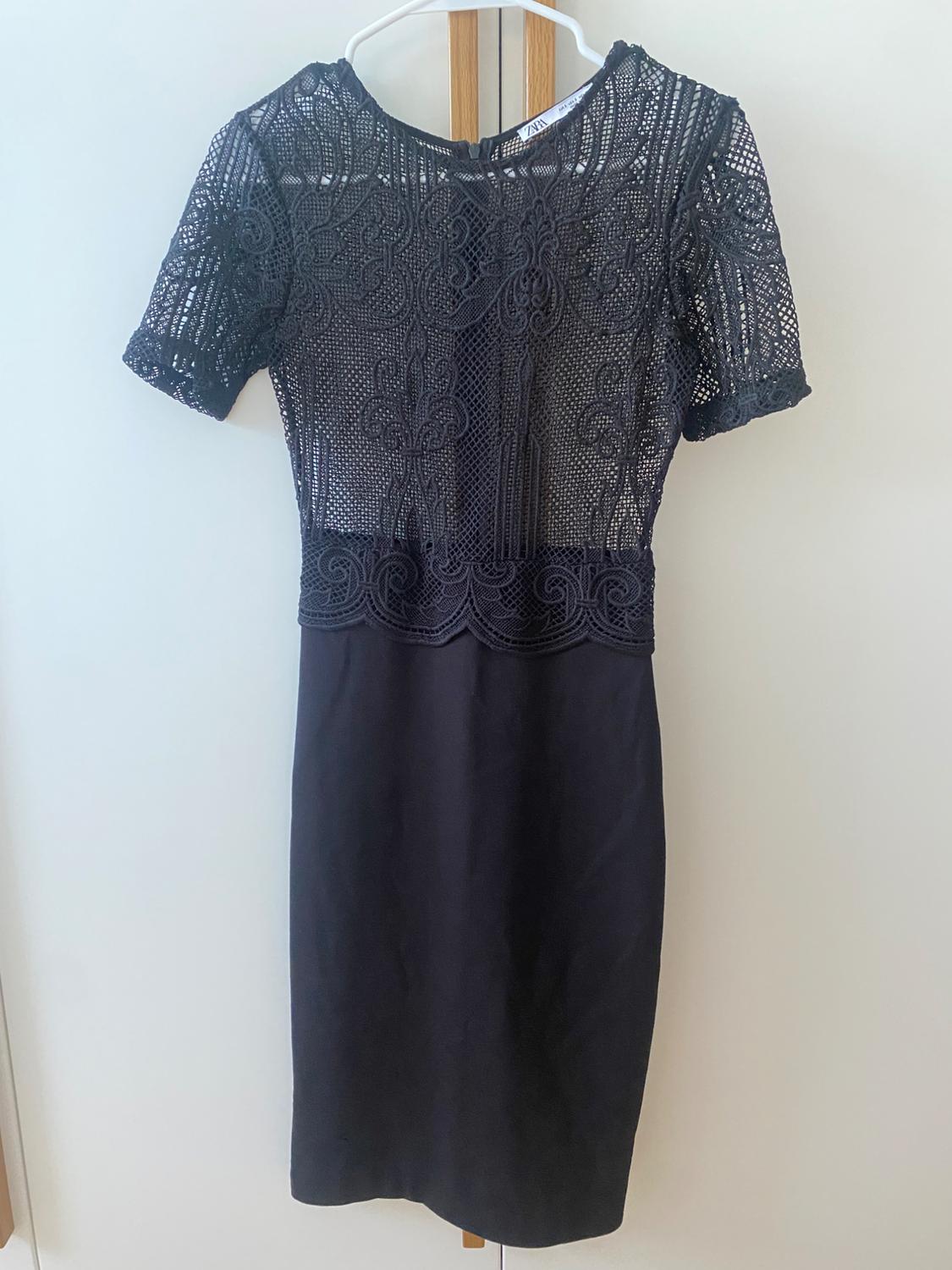 Black lace dress hanging on a white hanger over a door. Intricately designed top with short sleeves.