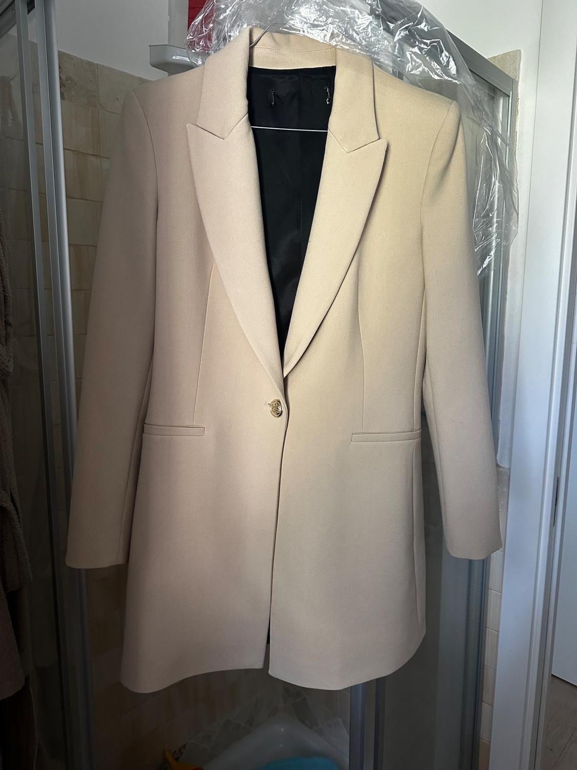 Beige coat hung on a hanger inside a shower, with a tiled background and transparent plastic curtain.