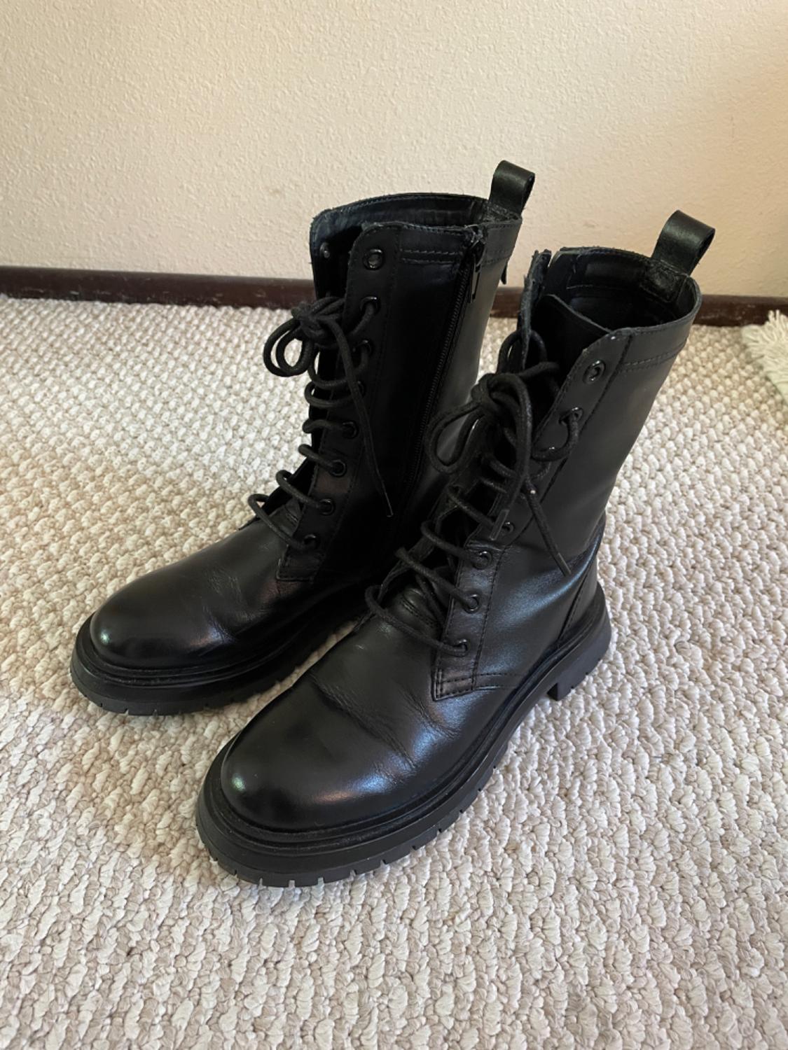 Black leather boots with laces and thick soles on a light rug, next to a beige wall.