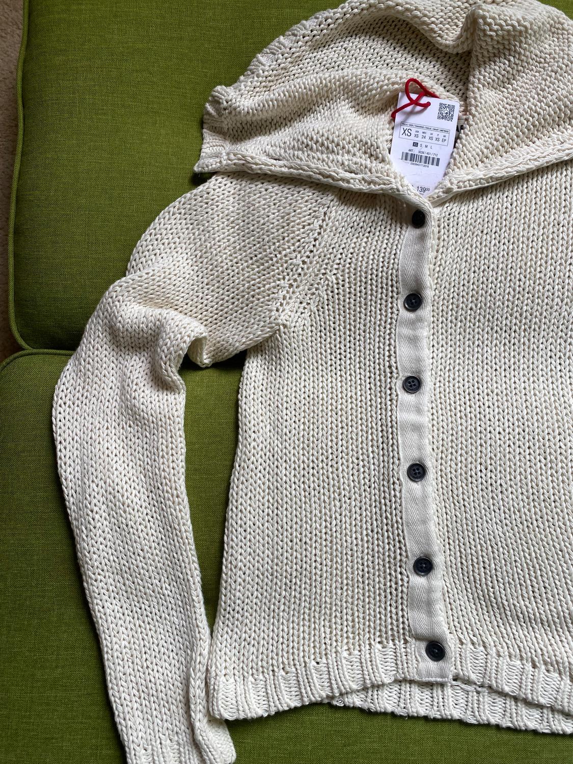 White knit cardigan with hood and black buttons on a green sofa. Visible tag with size XS and QR code.