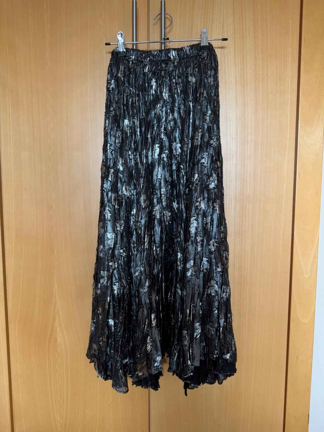 Long pleated skirt in dark tones with sparkling details, hanging on a hanger over a light wooden door.