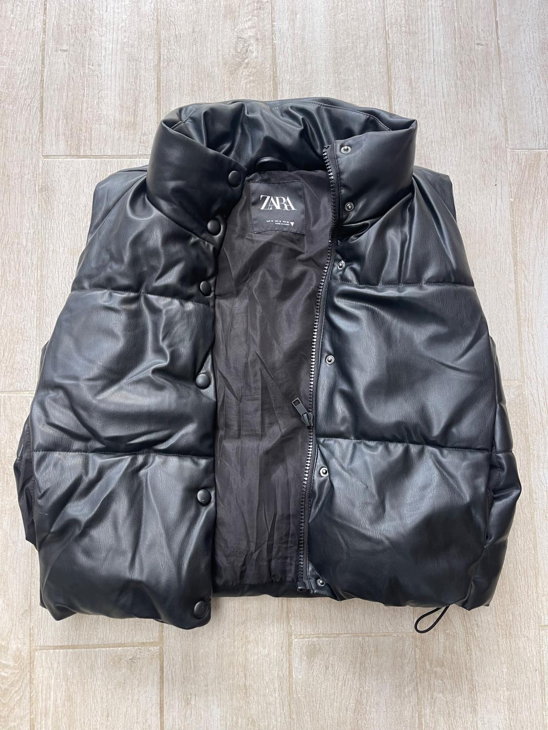 Black padded gilet with a glossy finish, zip-up fastening and snap button closure, visible inner label with Zara logo.