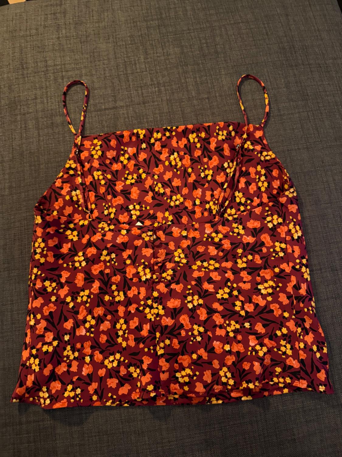 Camisole top with spaghetti straps in a floral print with shades of red, orange and yellow on a burgundy background, placed on grey fabric.