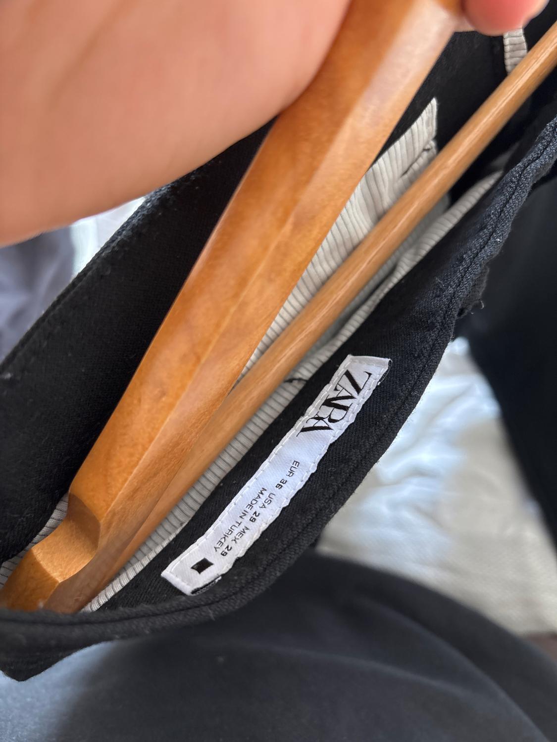 Hand holding a wooden hanger with a black garment. Visible label with text: ZARA EUR 36 USA 29 MEX 29 MADE IN TURKEY.