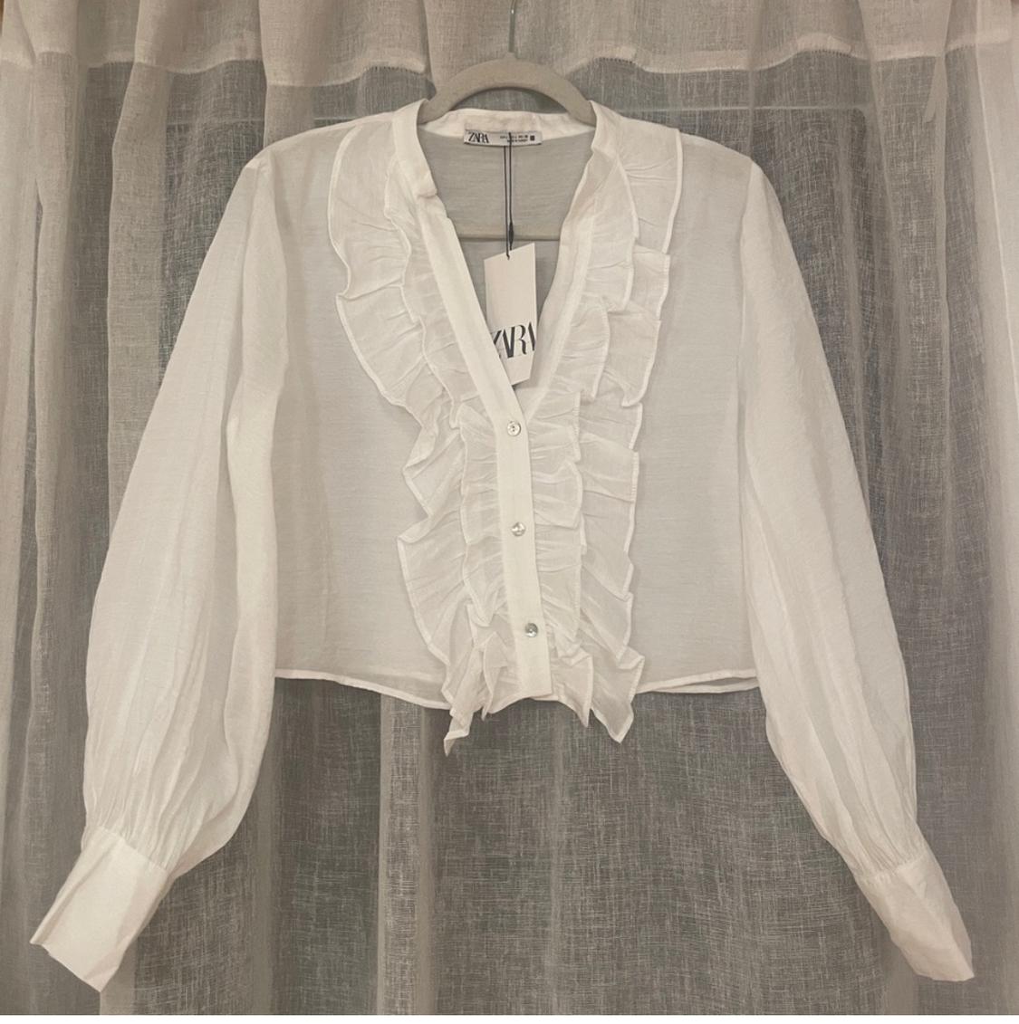 White blouse with ruffles and buttons, hanging on a hanger in front of a translucent curtain. Visible tag with text "ZARA".