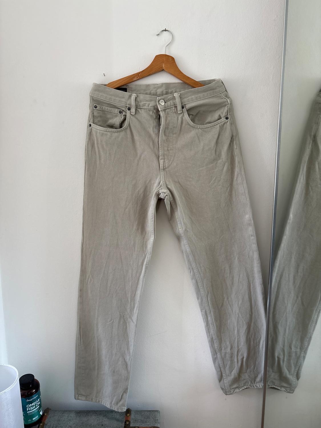 Beige jeans on a wooden hanger against a white wall. The edge of a mirror and a jar with the text OMEGA FISH OIL are visible.