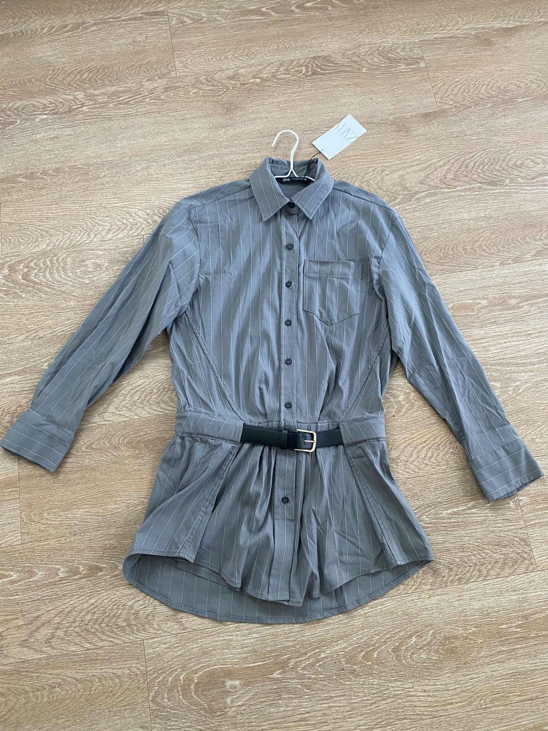 Fine-striped gray shirt with black belt, hanging on a hanger over a light wood floor. Visible tag.