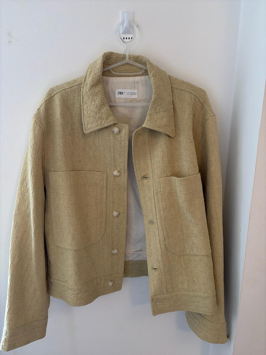 Beige textured casual-style jacket with buttons and pockets, hanging on a white wall. Interior label with the text ZARA.