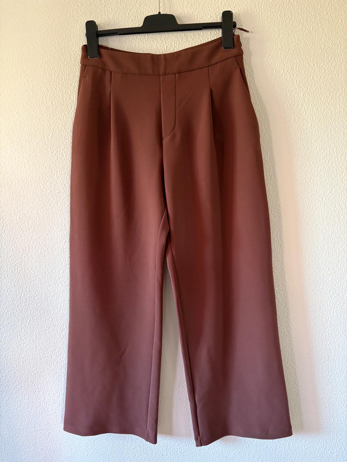 Brown wide-leg trousers with front pleats, hanging on a black hanger against a white wall.