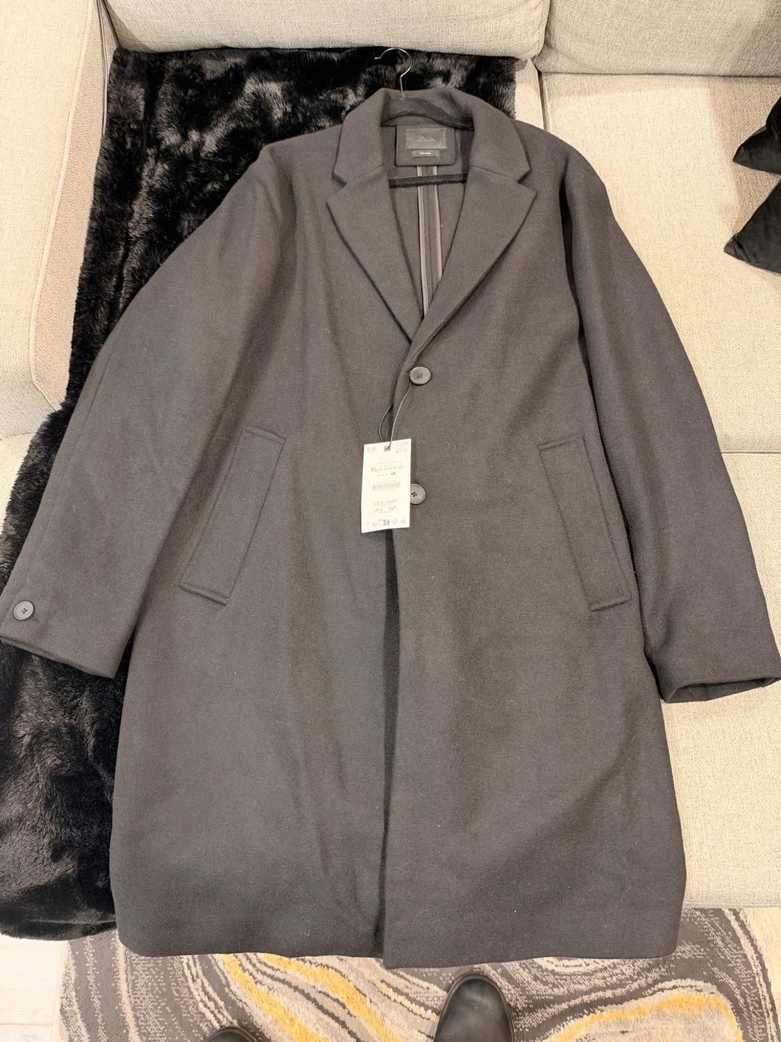 Long black coat ZARA with buttons and a price tag, hanging over a sofa with a faux-fur throw blanket.