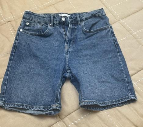 Blue denim shorts with pockets, laid flat on a beige quilted fabric with white stitching. Casual style.