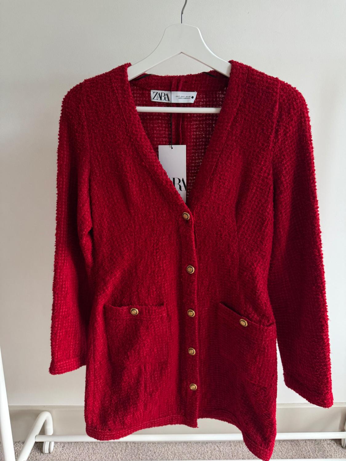 Red knitted cardigan with gold buttons and front pockets, hanging on a white hanger. Visible label with Zara logo.