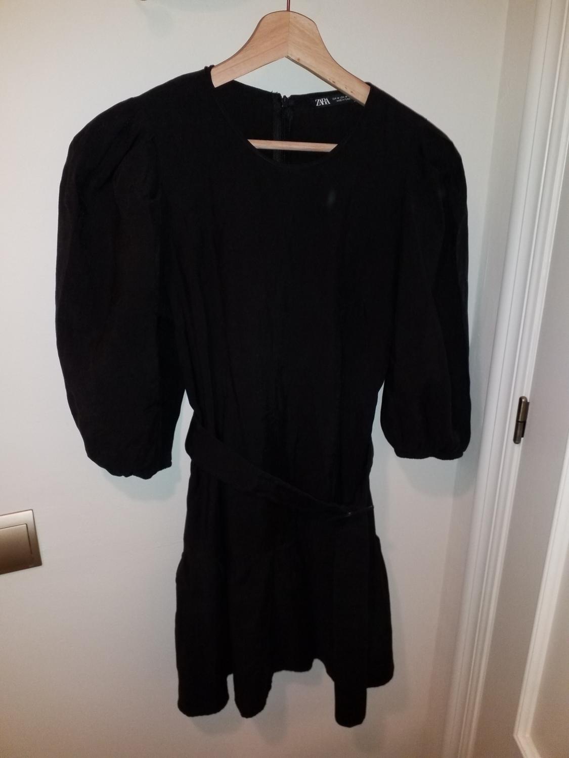 Black puff-sleeved dress hanging on a wooden hanger. Visible label with the ZARA brand. White wall background.