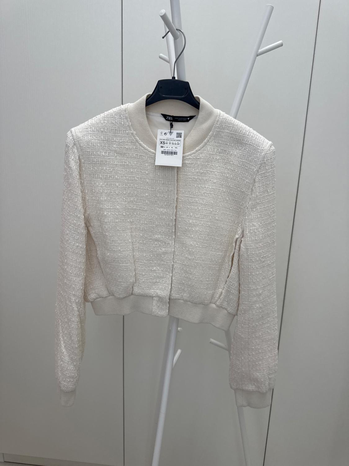 White textured fabric jacket hanging on a white clothes rail. Visible label with size XS and barcode. Neutral background.