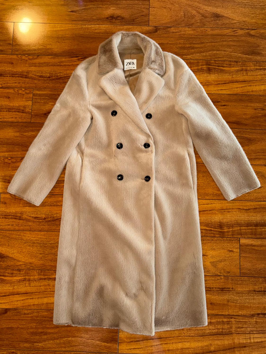 Long beige coat with black double-breasted buttons on a wooden floor. Visible label with text "ZARA".