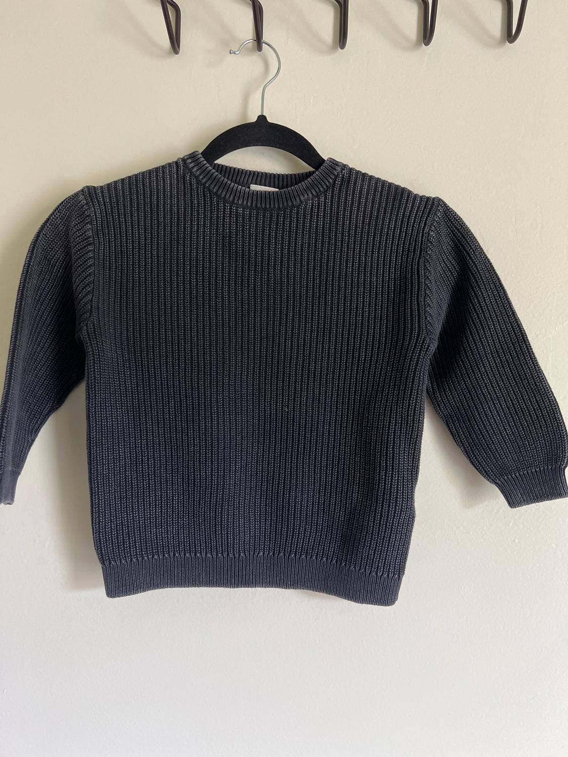 Dark grey knitted jumper hanging on a black hanger against a white wall. Round neck and long sleeve design.