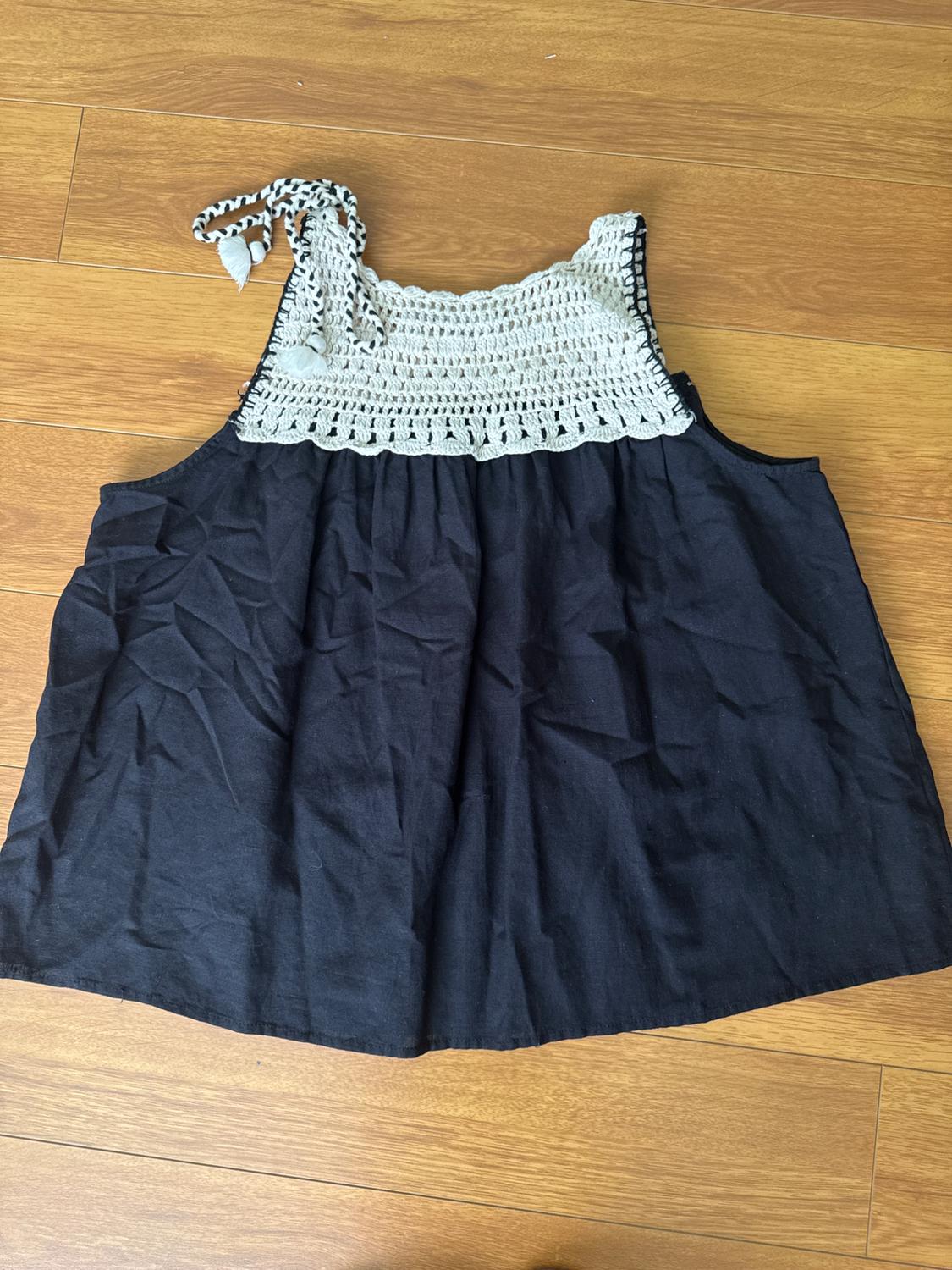 Black sleeveless blouse with white crochet top and adjustable straps, on a light wooden floor.