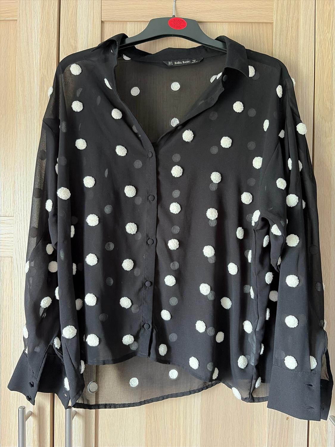Black semi-sheer shirt with raised white polka dots, hanging against a wooden background. Text: ZARA BASIC, 14 UK/IRL, 42 EUR, 10 USA.