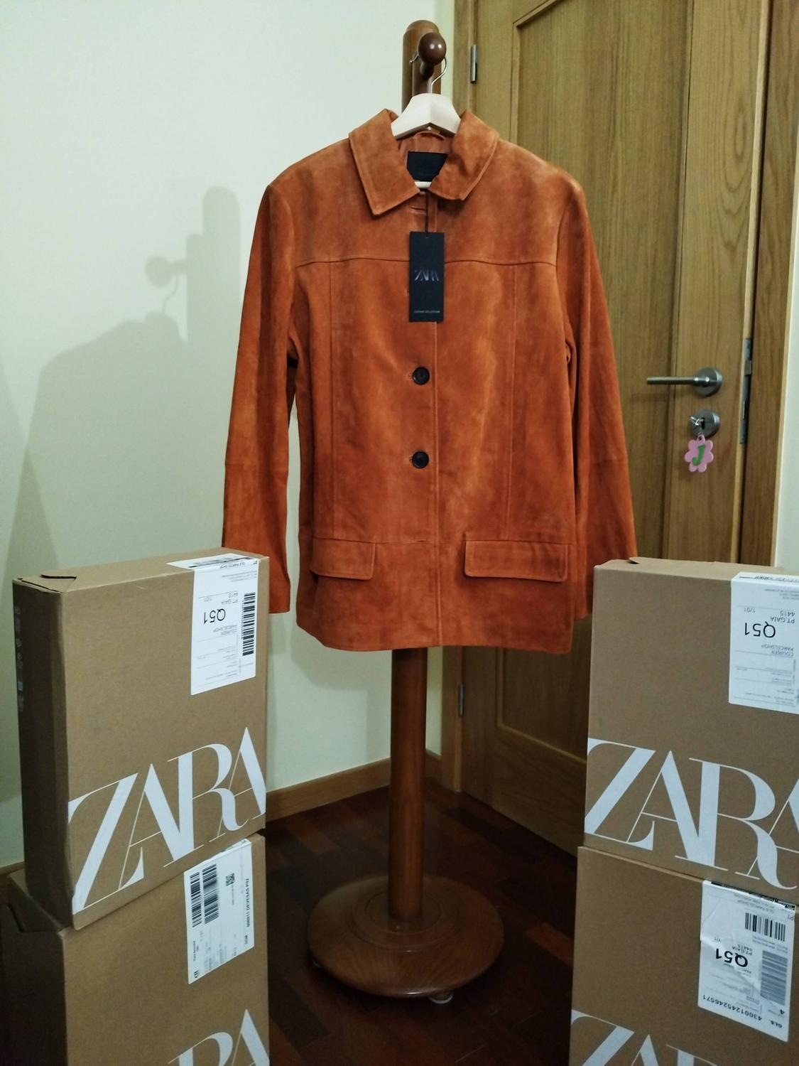 Brown jacket on a clothes rack surrounded by cardboard boxes with the Zara logo, in a room with a wooden door.