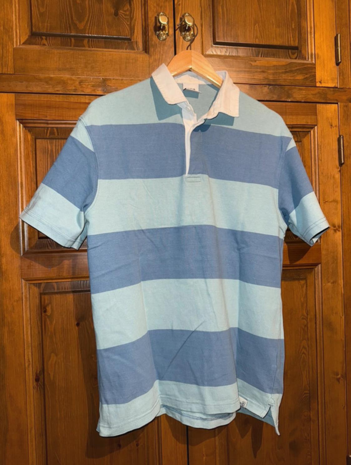Blue and sky-blue horizontal striped polo shirt hanging on a wooden hanger, in front of a brown wooden wardrobe.