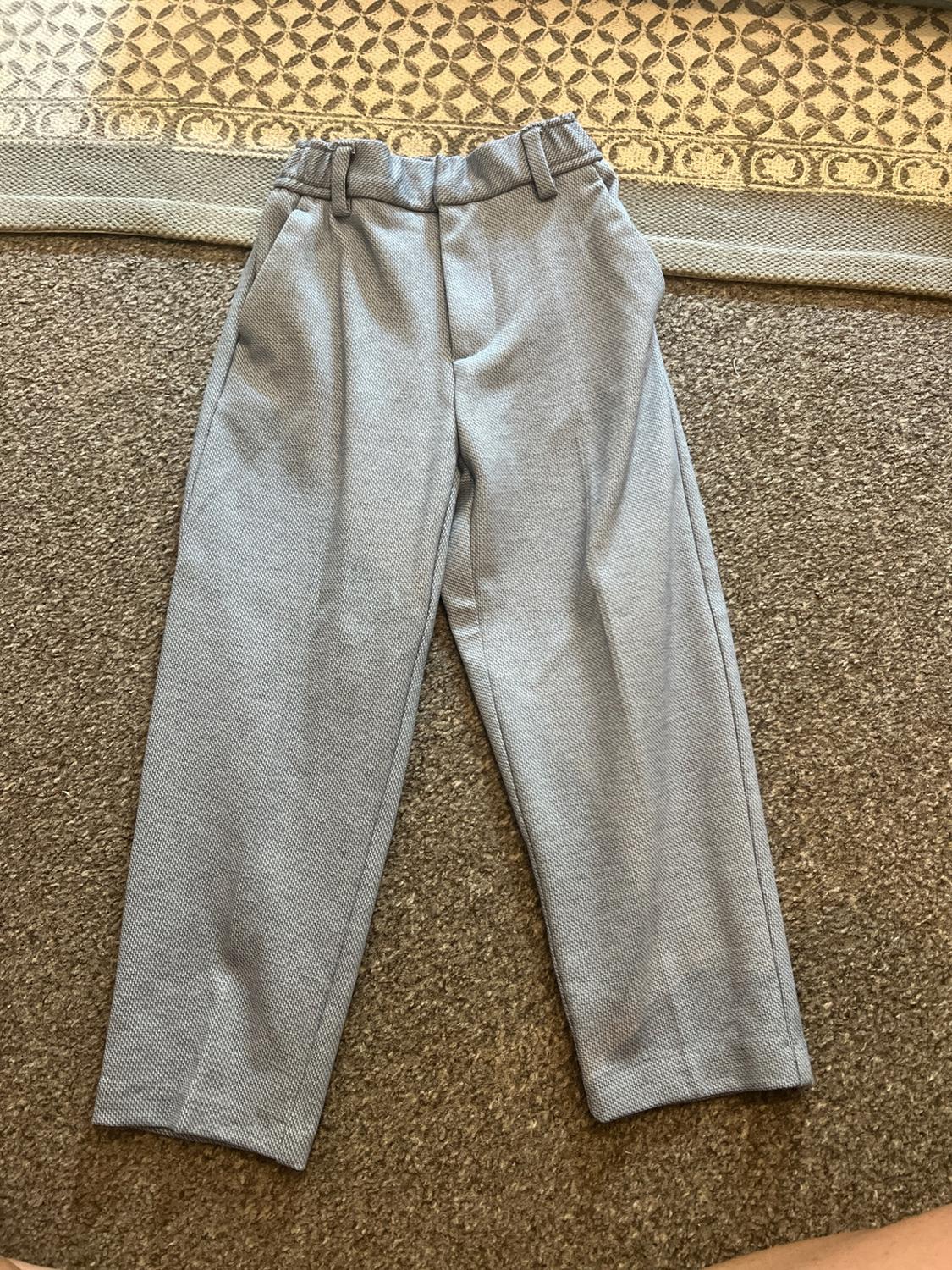 Light grey textured trousers with an elasticated waistband and belt loops, laid out on a brown rug and another with a geometric pattern.