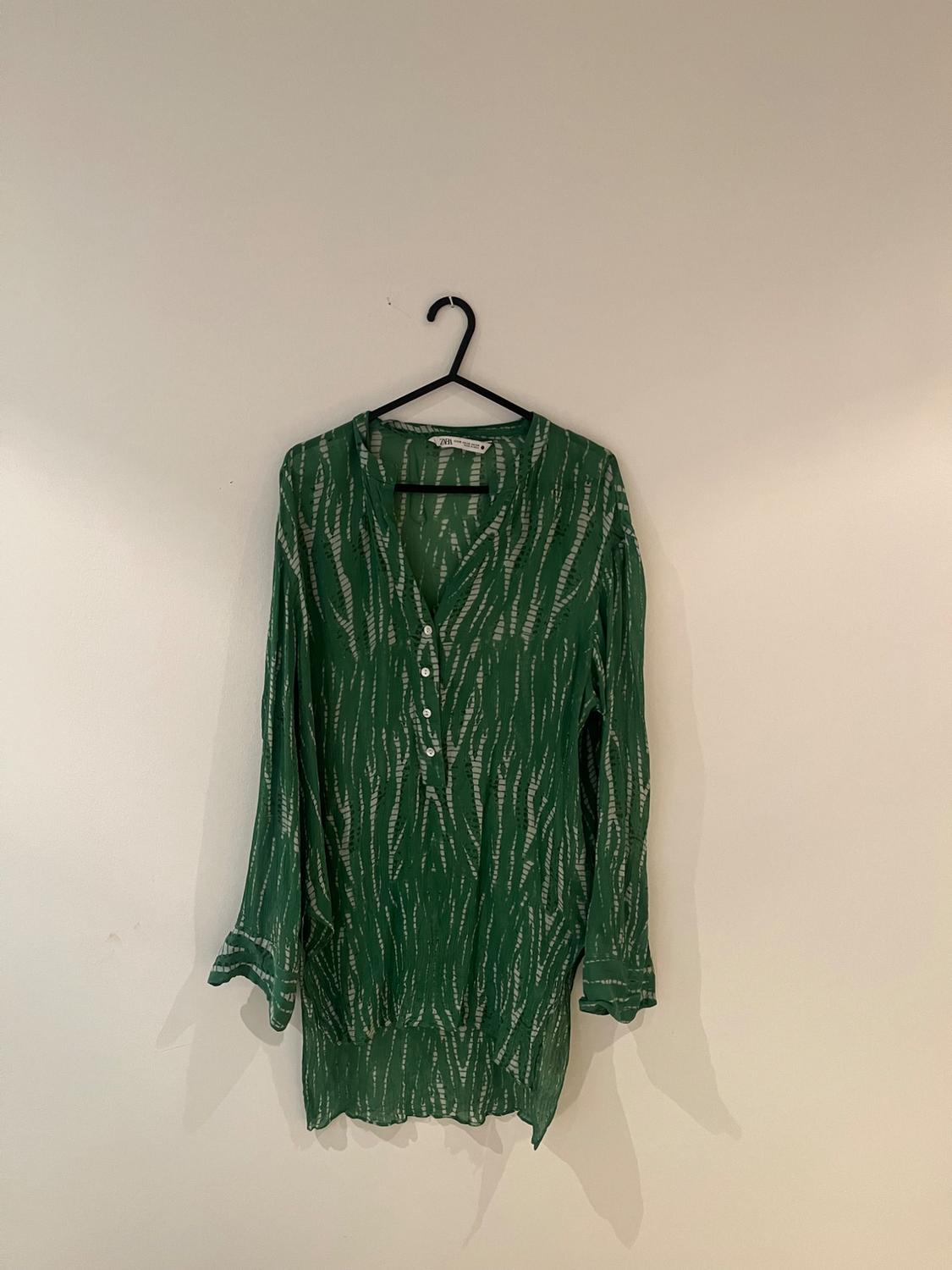 Green blouse with a white pattern hanging on a black hanger against a white wall.