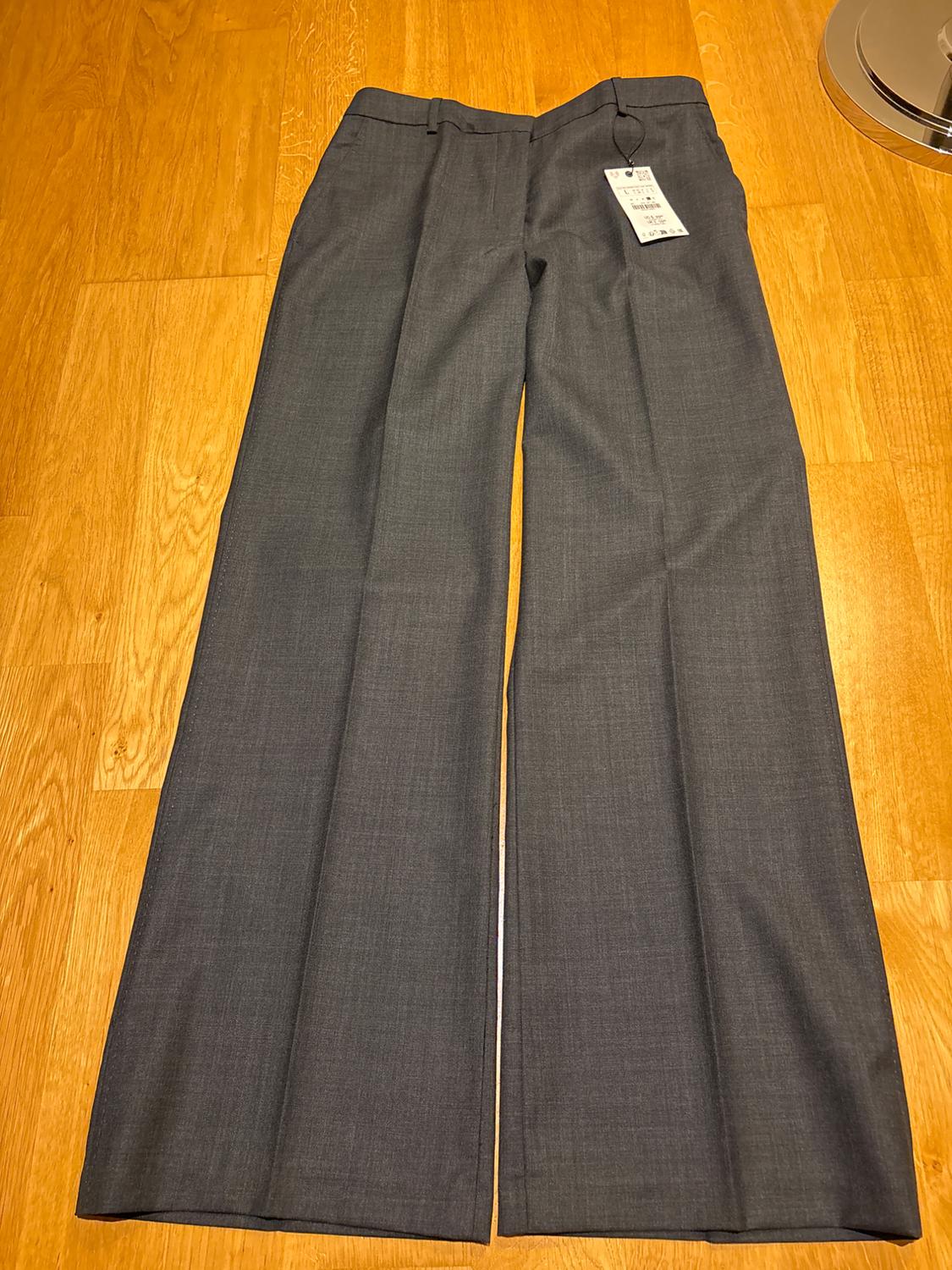 Grey wide-leg trousers on a light wooden floor, with a visible hang tag at the waist.