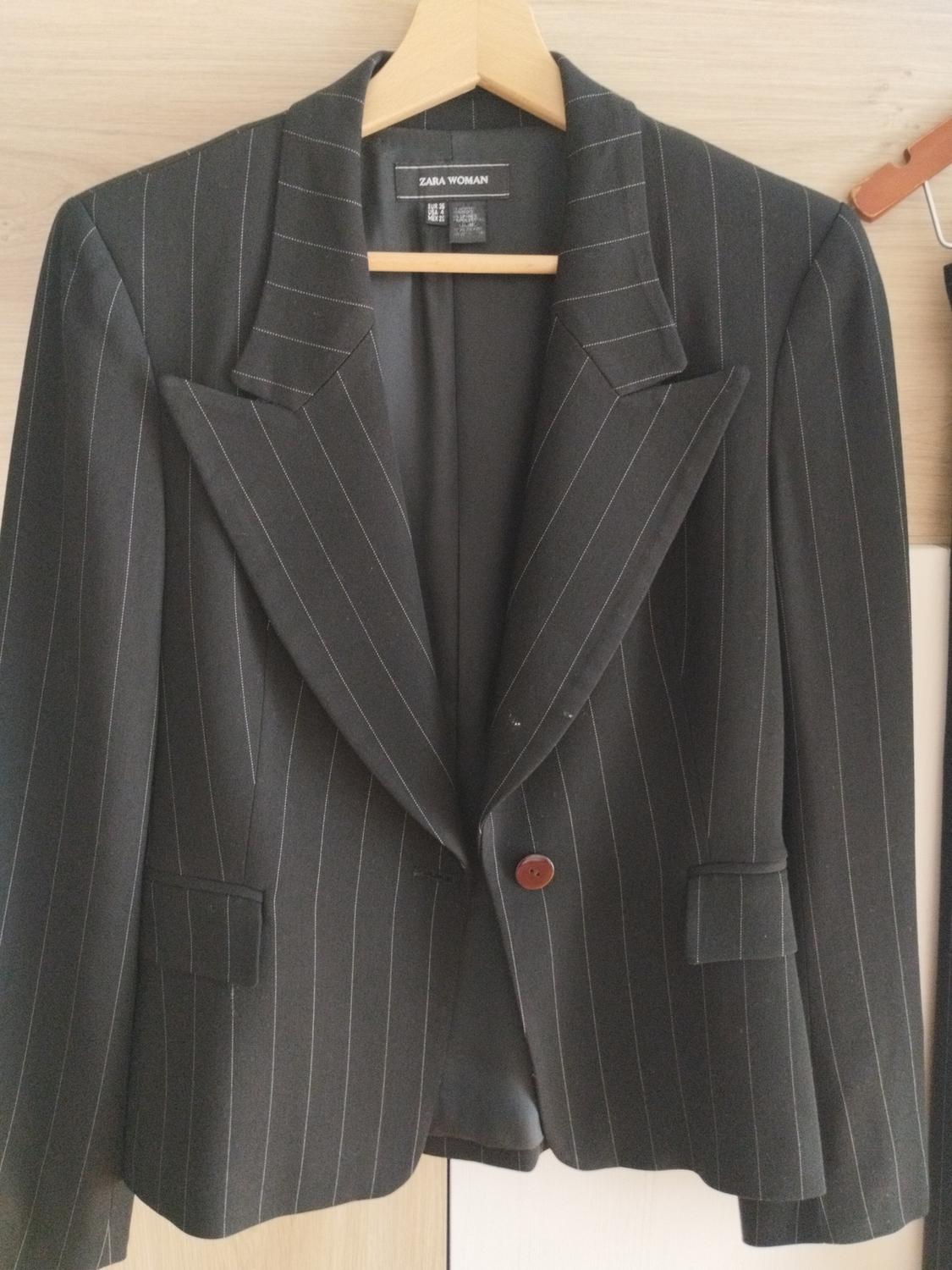 Black pinstripe blazer on a wooden hanger. Peak lapels, one button and pockets. Inner label with the text ZARA WOMAN.