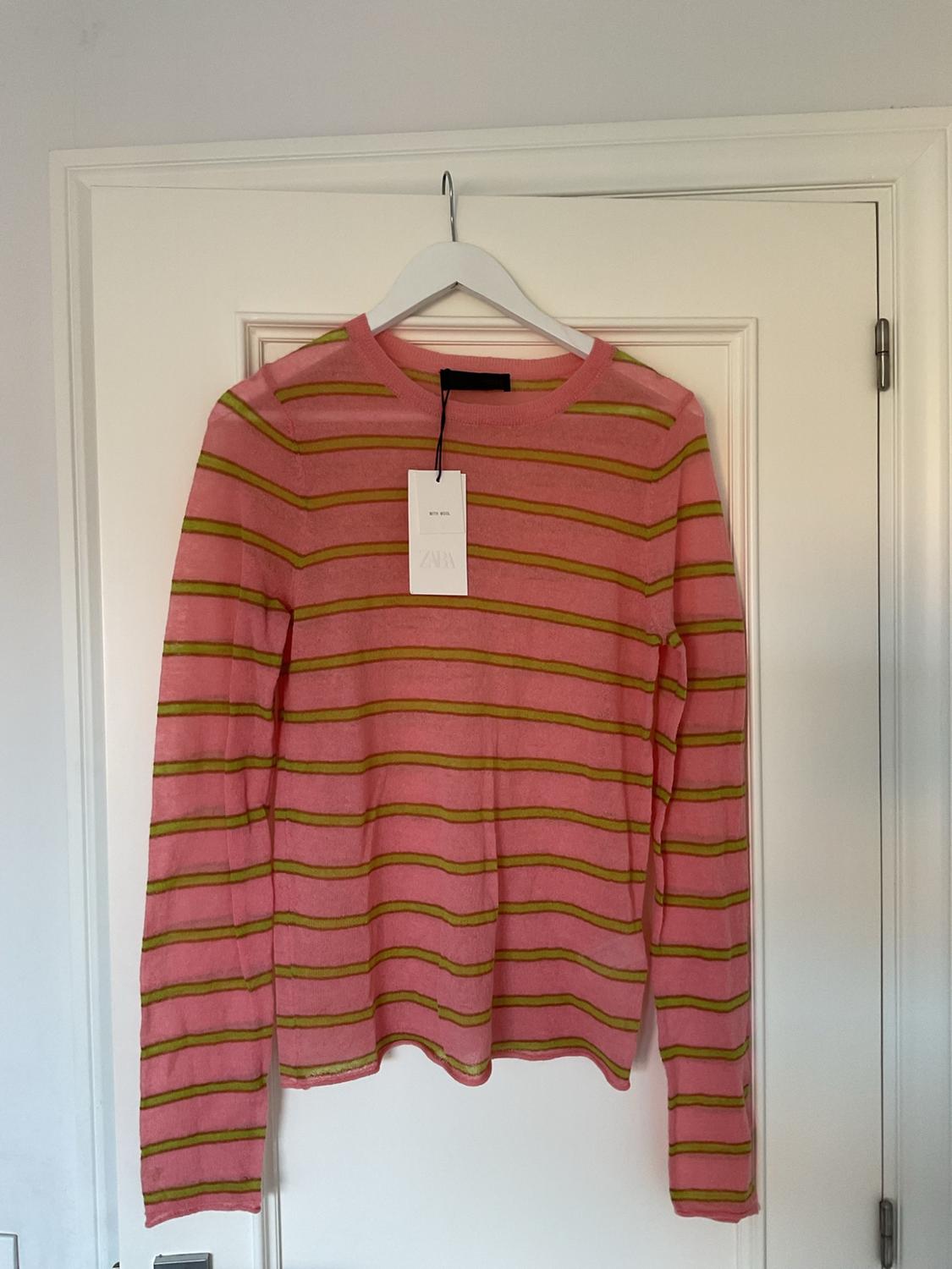 Pink long-sleeved jumper with horizontal green stripes hanging on a white door. Label with the text WITH WOOL ZARA.