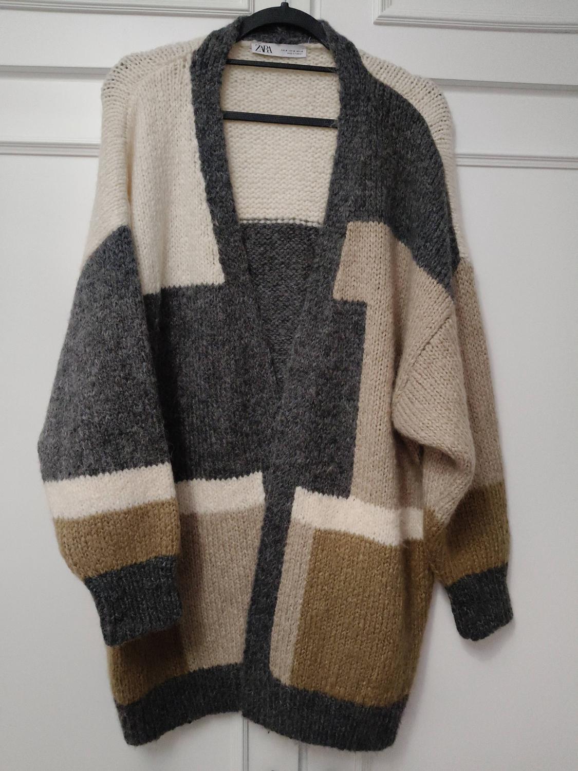 Chunky knit cardigan with geometric design in beige, grey, and brown tones, hanging on a black hanger in front of a white door.