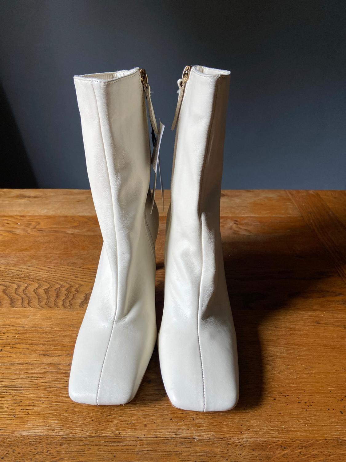 Pair of white square-toe boots with centre stitching, featuring zip tags, on a wooden table against a dark background.