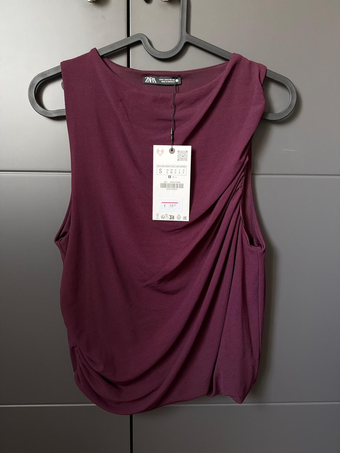Burgundy sleeveless blouse hanging on a black hanger, with a price tag of €19.95 visible in the centre.