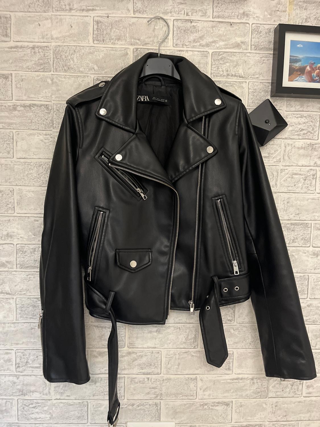 Black faux leather jacket with silver zips and buttons, hanging on a white brick wall. Visible text: "ZARA".