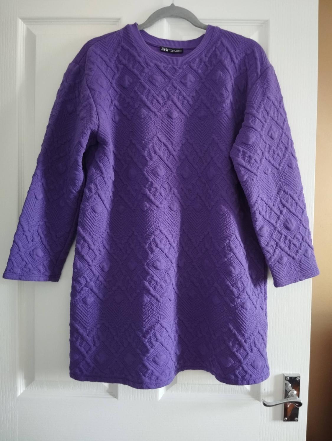 Long-sleeved purple dress with a geometric texture, hanging on a hanger against a white door. Label with the text ZARA on the neck.