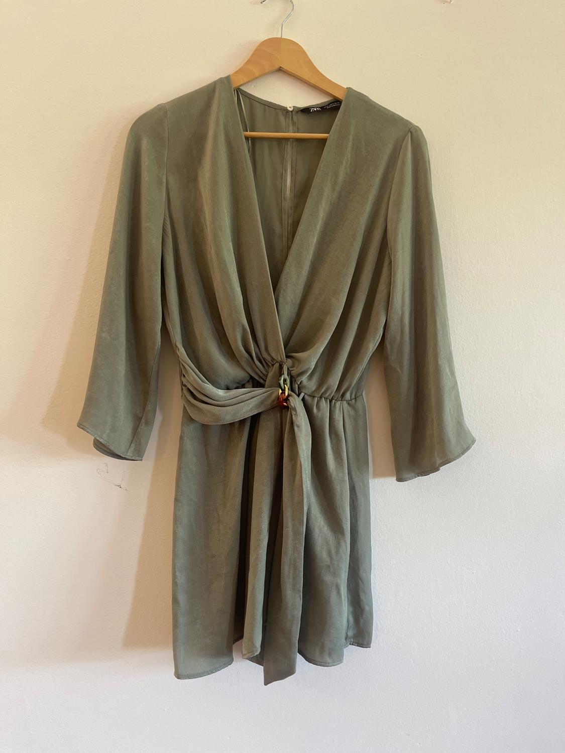 ZARA olive green short dress with a V-neck, 3/4 sleeves, and a front knotted detail, hanging on a wooden hanger against a white wall.