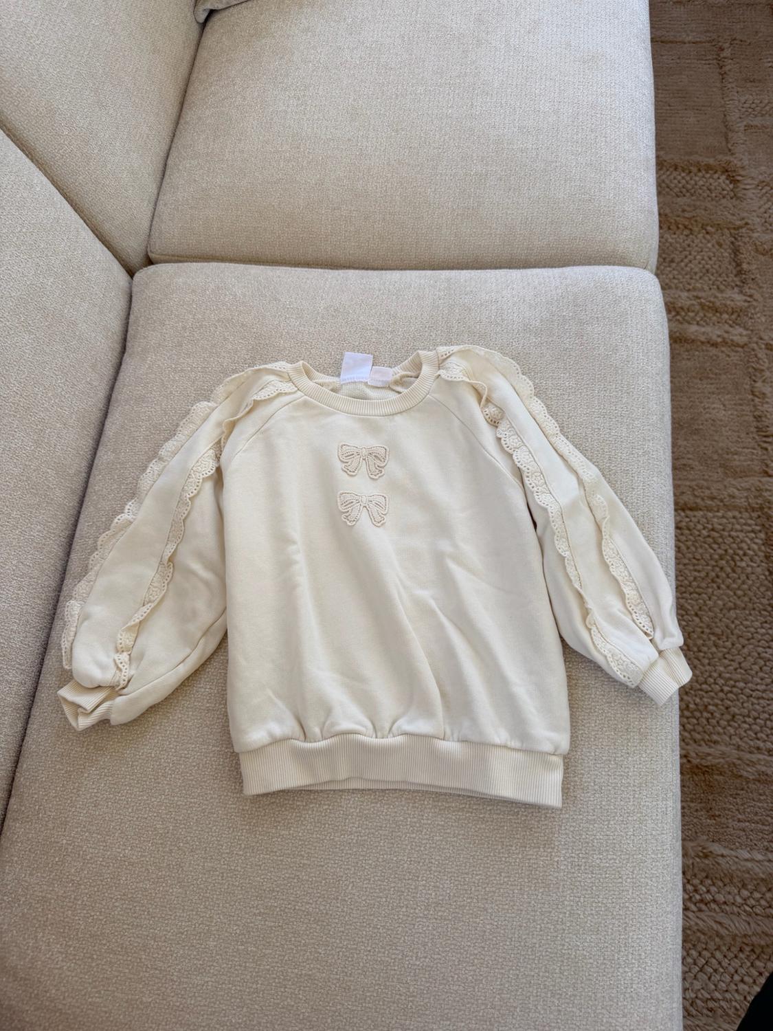 Cream-colored hoodie with two embroidered front bows and lace ruffles on the sleeves, on a light-colored sofa.