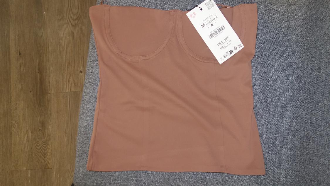 Light brown sleeveless top on a gray background, with a white label indicating size M, US S, UK 8. Wooden surface visible.