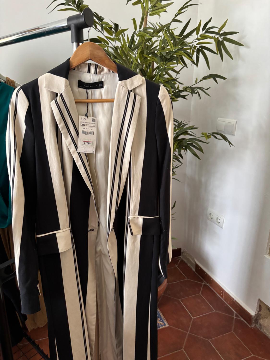 Black and white striped long coat on a coat rack. Interior with a plant. Visible text: ZARA WOMAN XS € 79.