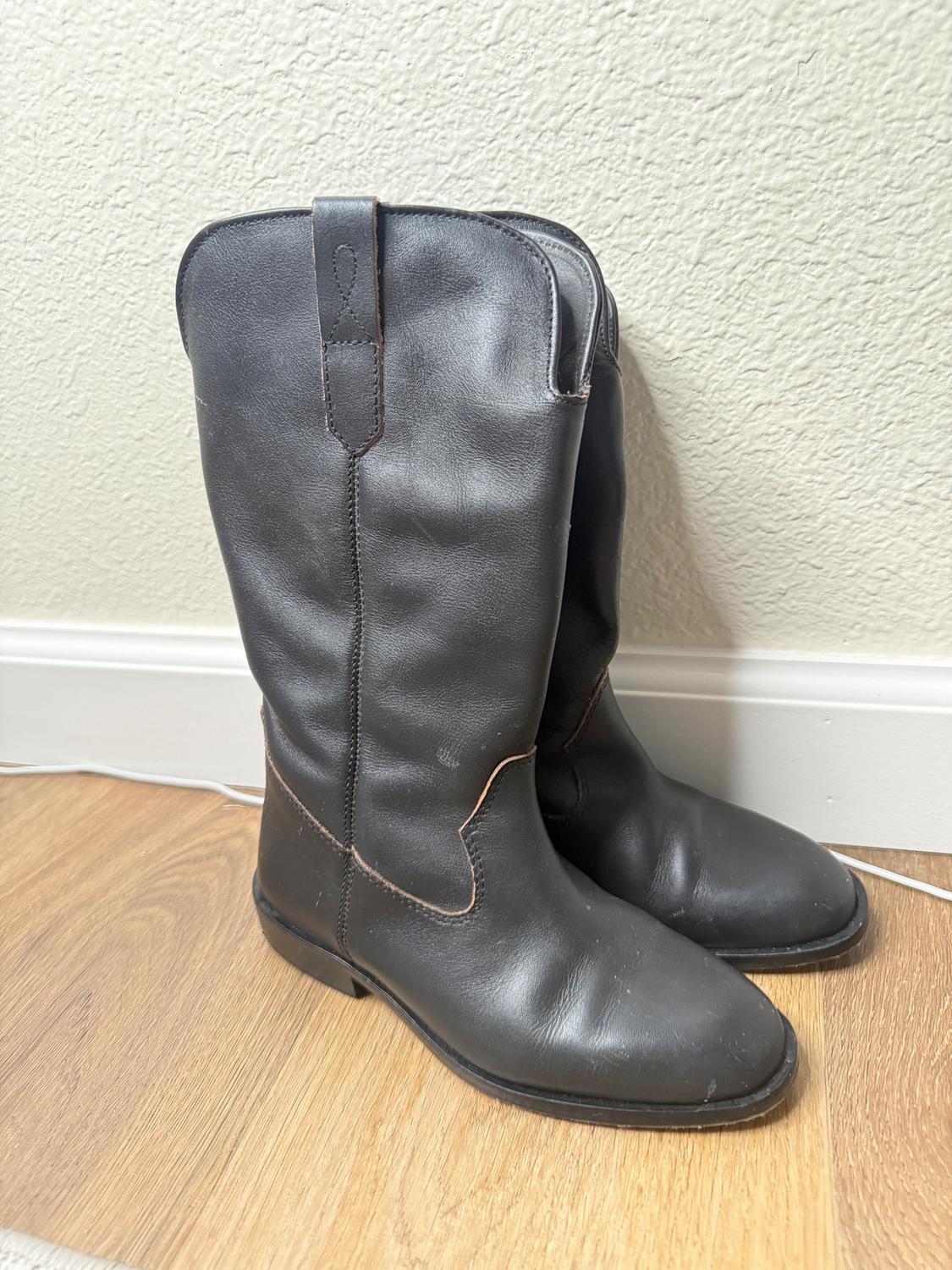 Black leather boots on a light wooden floor, leaning against a textured white wall.