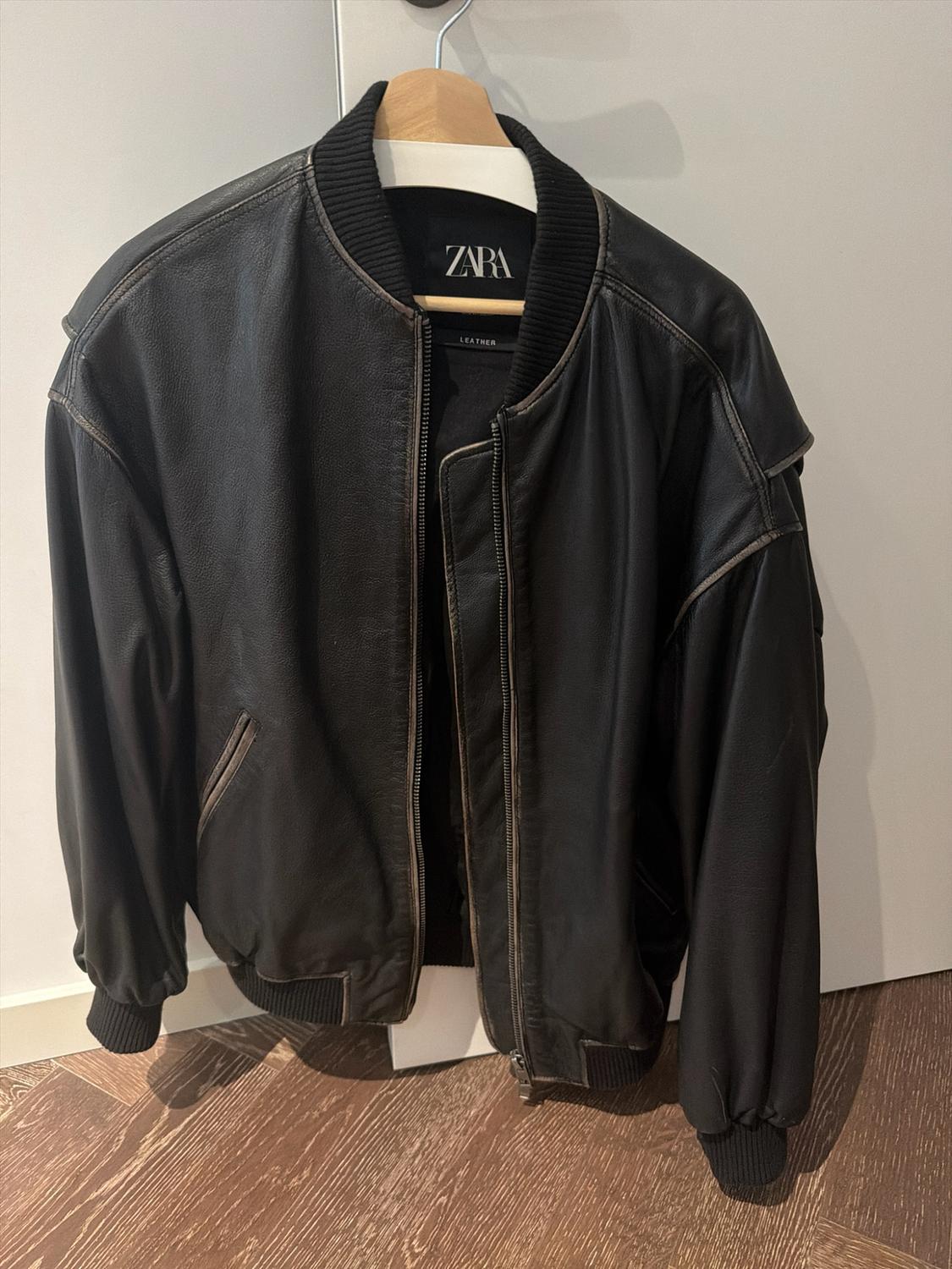 Black leather bomber jacket with a zip hanging on a hanger. Inner label with the text ZARA LEATHER.