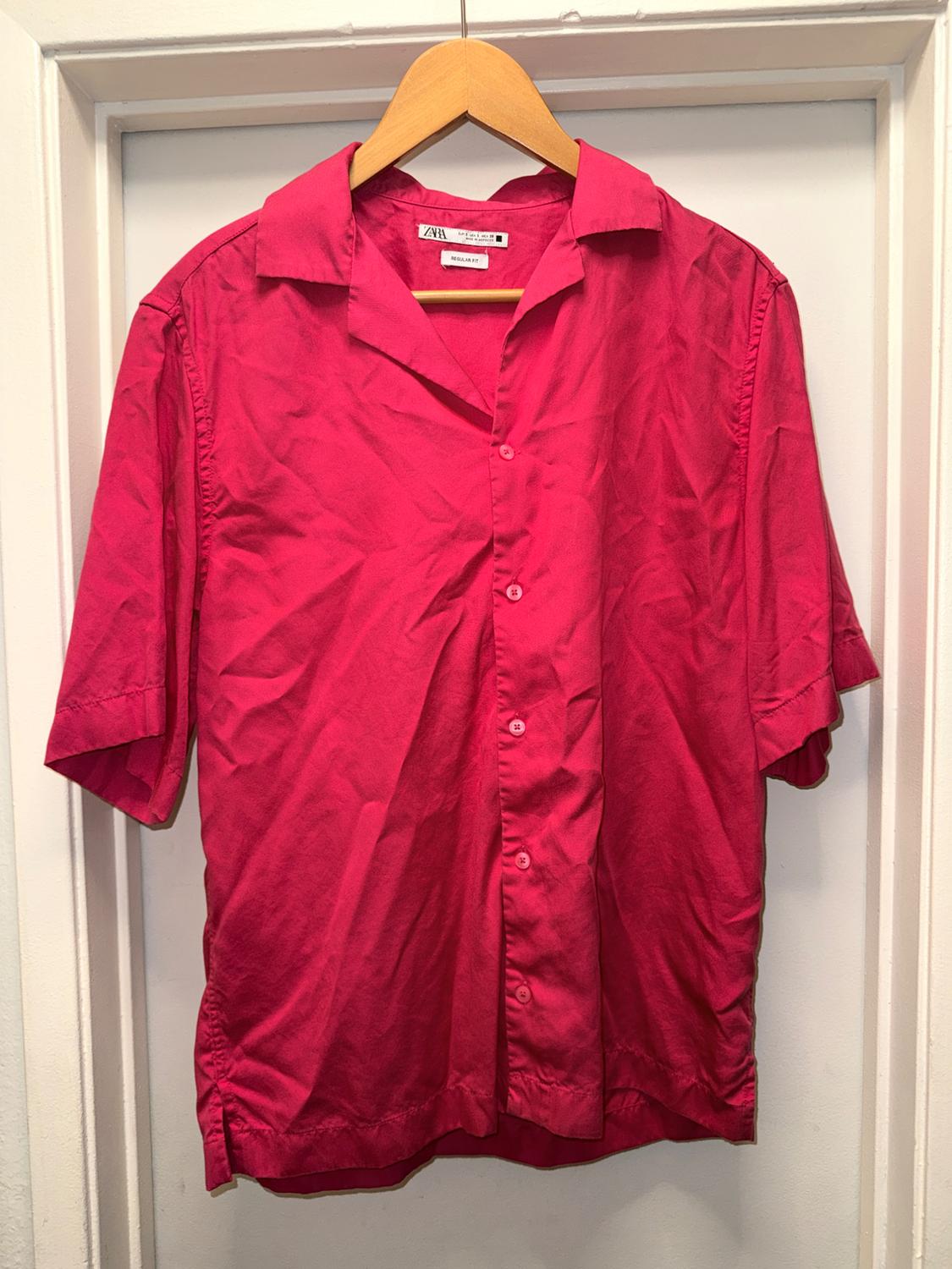 Hot pink short-sleeved button-down shirt, hanging on a wooden hanger over a white door. Label with the text ZARA.