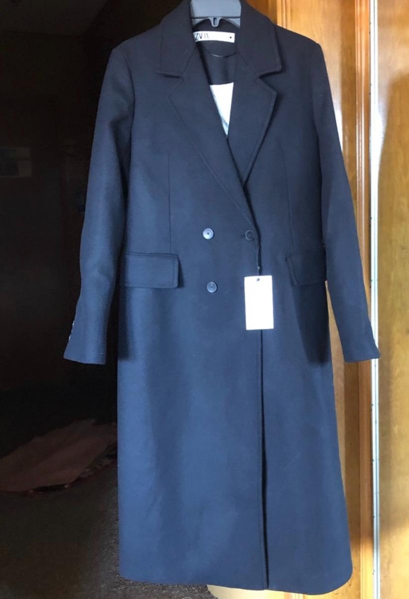 Long black coat hanging on a hanger, with wide lapels and double buttons. White label visible on the front. Wooden background.