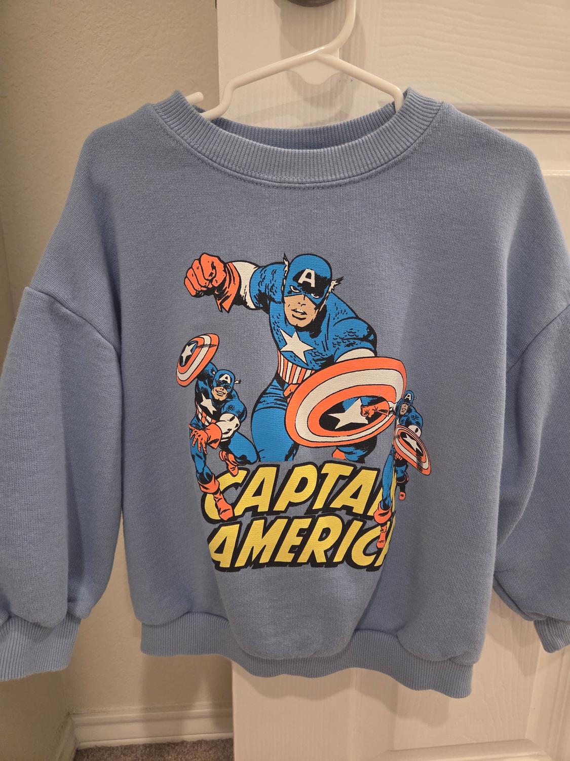 Blue sweatshirt with Captain America print on the front, hanging on a white hanger in front of a white door.