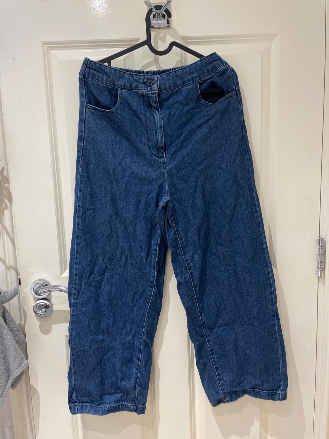 Loose-fitting blue jeans with gathered hems, hanging on a black hanger on a white door with a silver handle.