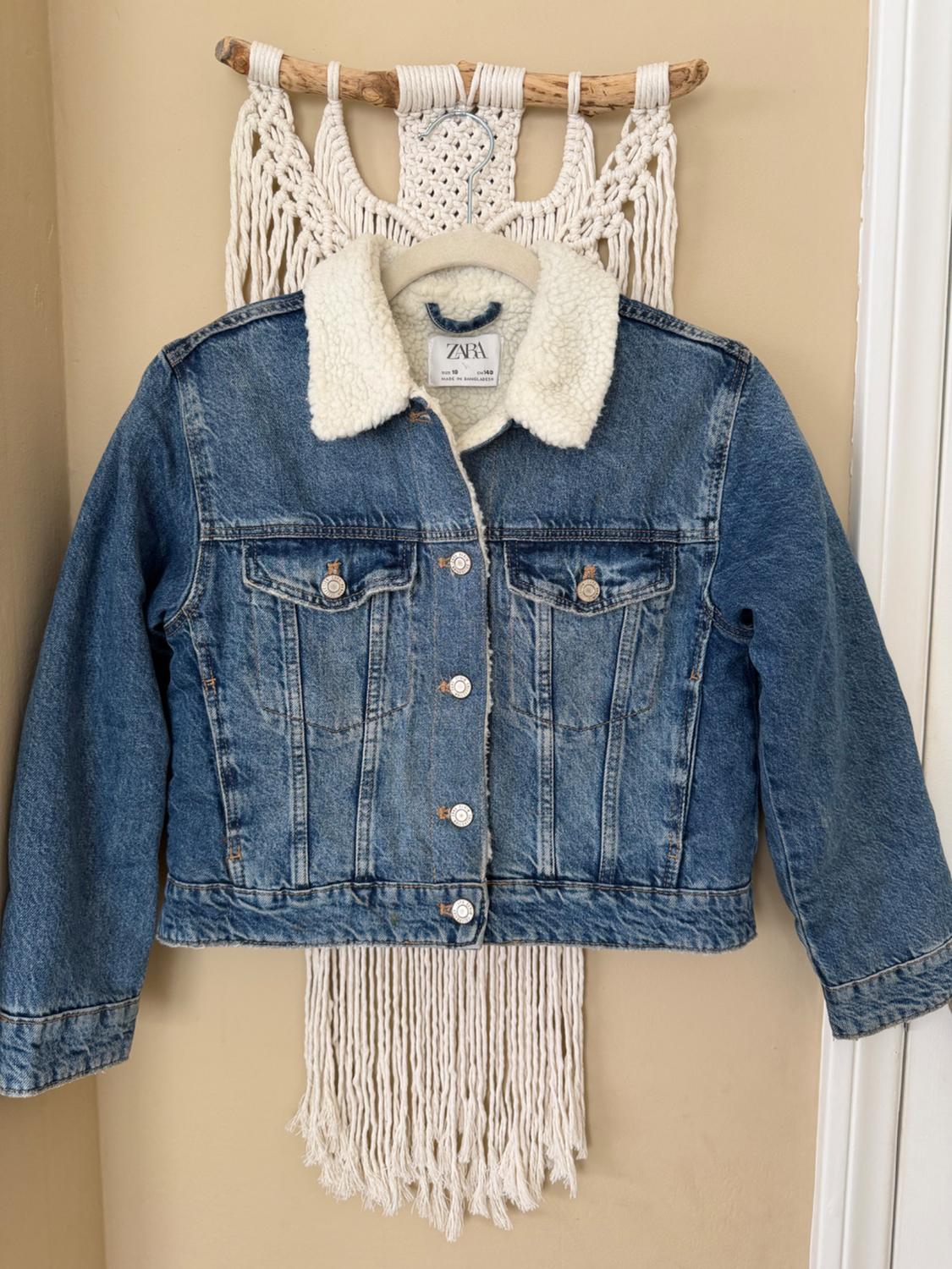 A blue denim jacket with a wool collar is hanging on a hanger over a white macrame tapestry. The visible tag reads: ZARA.