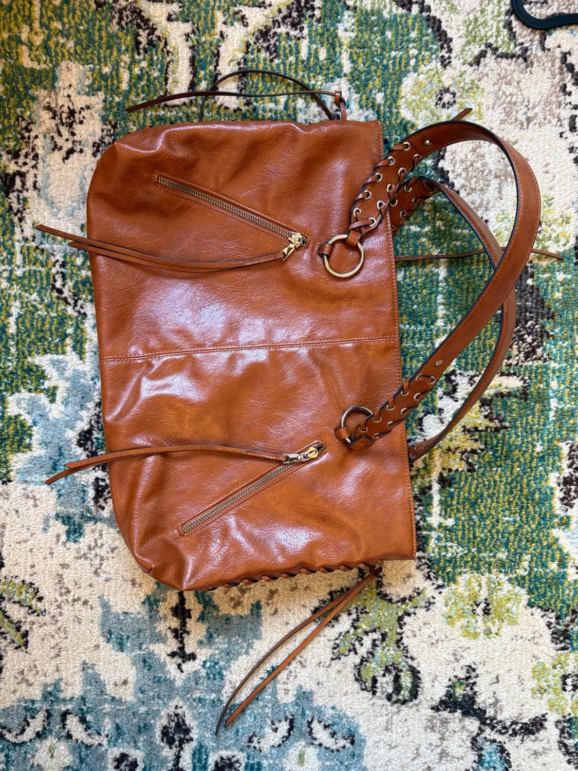 Brown handbag with diagonal zips and braided handles, resting on a patterned rug in shades of green, blue and beige.
