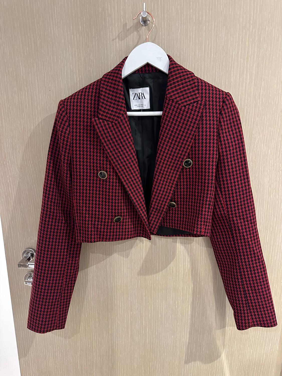 Red and black houndstooth double-breasted cropped jacket, hanging on a hanger over a wooden door. Label with the text ZARA.