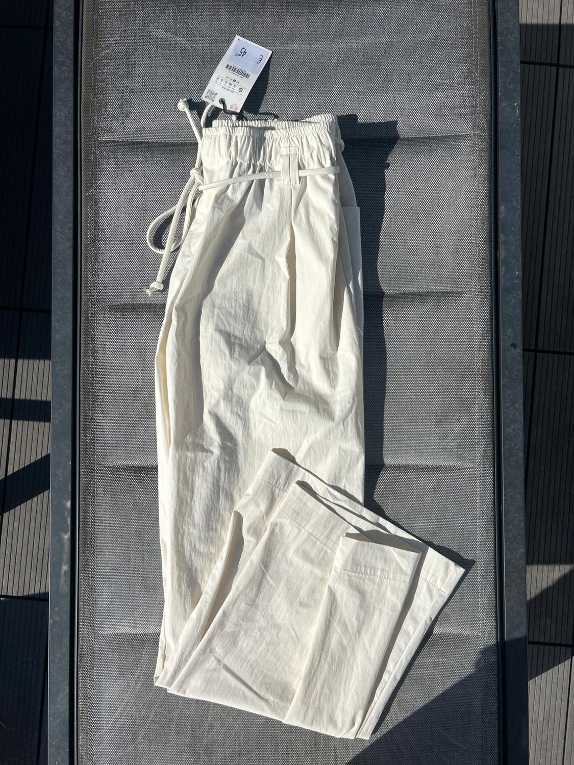 Ecru trousers with an elasticated waist and drawstring, folded on a grey sun lounger in the sun. Tag displaying the text "€ 45" and "S".